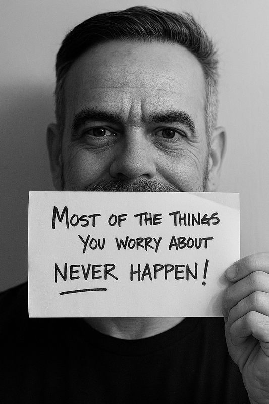 Black-and-white photo of a middle-aged man holding a sign in front of his mouth that reads, “Most of the things you worry about never happen!”