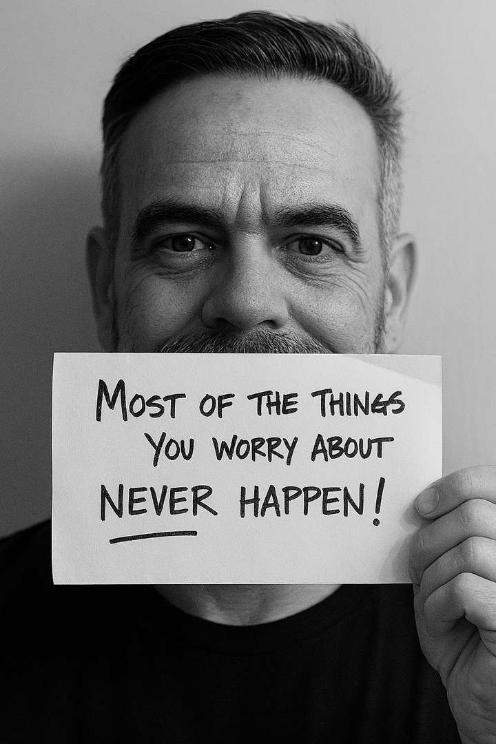 Black-and-white photo of a middle-aged man holding a sign in front of his mouth that reads, “Most of the things you worry about never happen!”