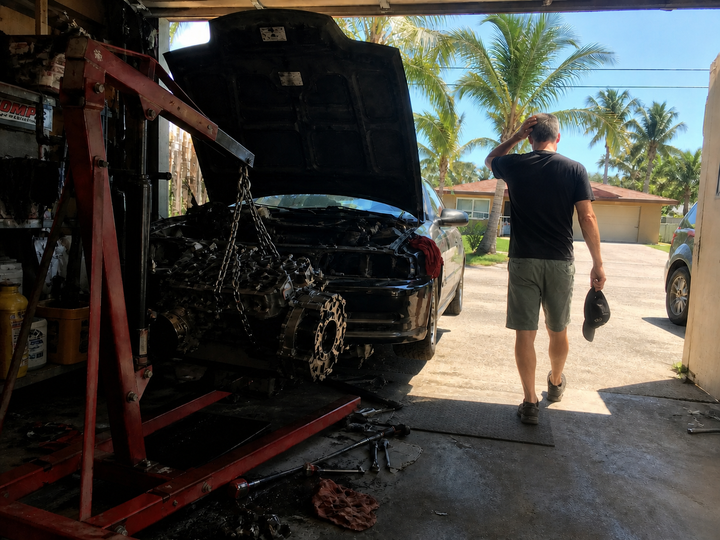 Man walking away into a sunlit driveway from an unfinished car engine repair in an open garage, stepping back to clear his mind