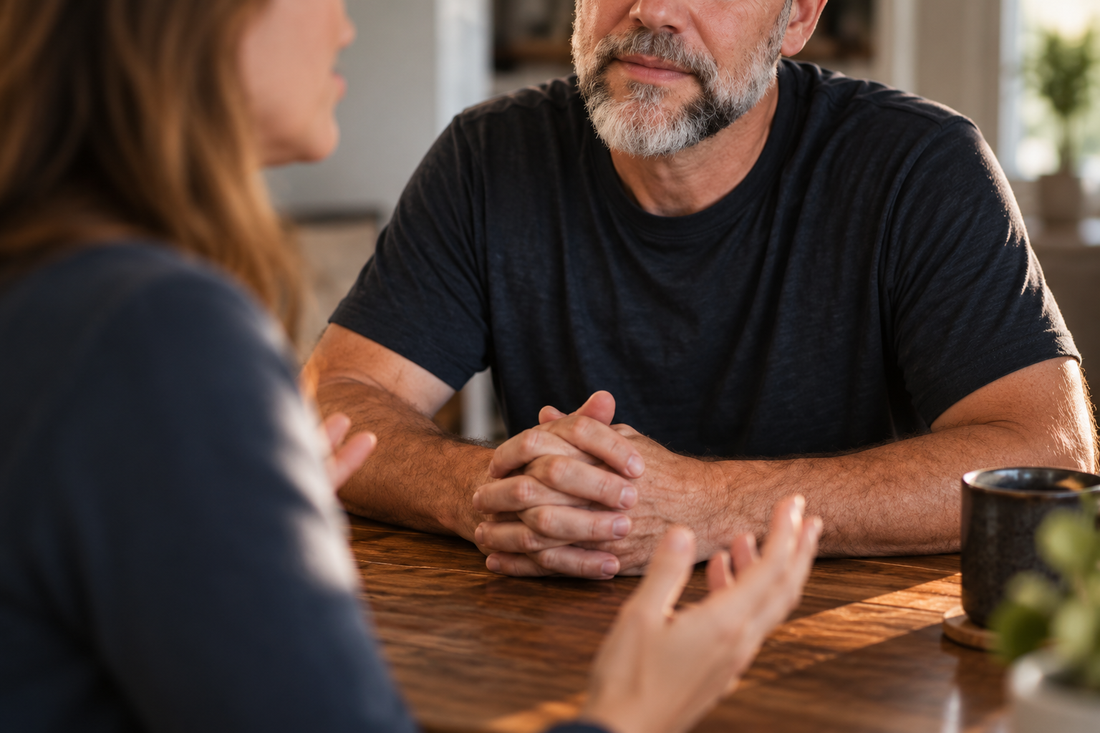 A man listens thoughtfully with hands clasped while a woman speaks across a wooden table.